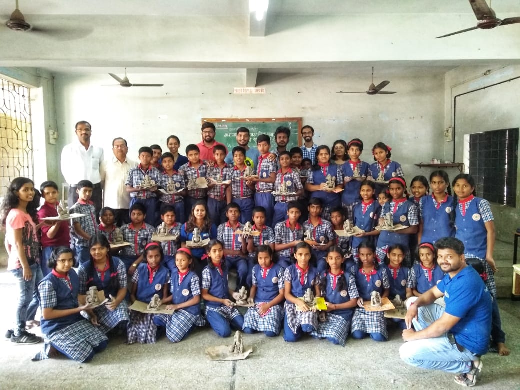 Approach volunteers with schoolchildren in blue uniforms, each holding a small clay figure they made in a classroom