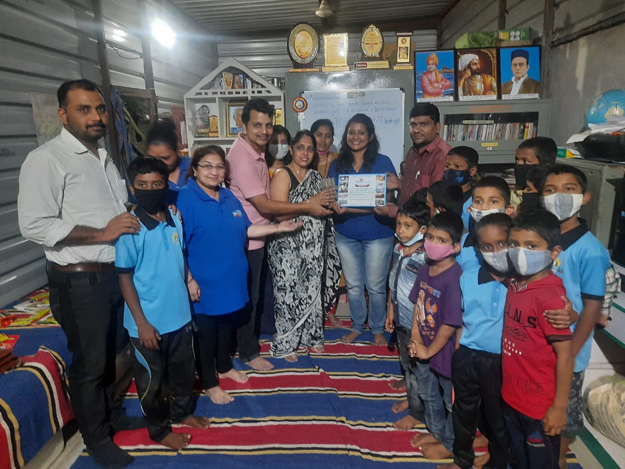 Volunteers and children wearing masks posing together in a small room with shelves and a colourful rug.